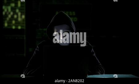 Man in mask cracking codes on laptop, falsification in elections, vote rigging Stock Photo