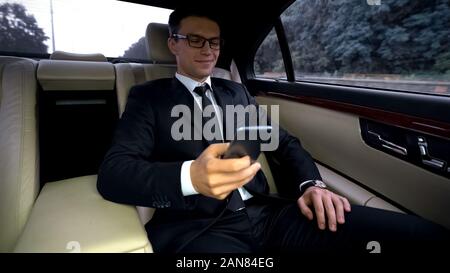 Delighted businessman riding on backseat of expensive car, using phone app Stock Photo