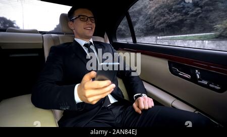 Smiling handsome businessman riding in luxury car, reading good news on phone Stock Photo