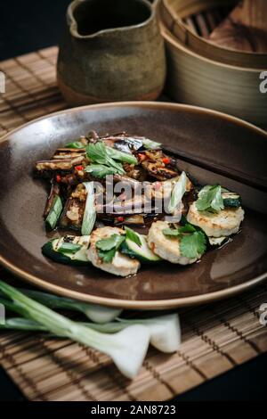 Vegan food. Asian spicy aubergine main with garlic, tofu, chilli and spring onions served on a plate with a bamboo matt and chopsticks. Stock Photo