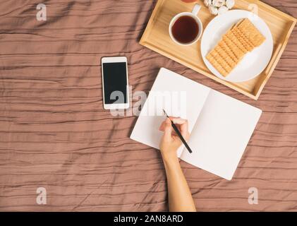 woman writes in large white open notebook, lay on bed with smartphone, breakfast tray, top view, copy space Stock Photo
