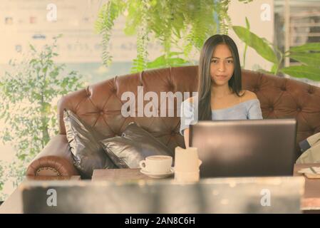 Young beautiful Asian teenage girl with laptop and cappuccino on wooden table while sitting at outdoor coffee shop Stock Photo