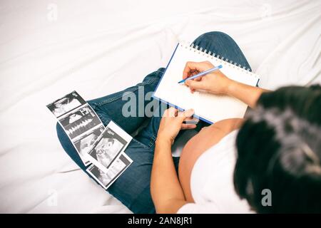 Pregnancy dairy. Pregnant woman writing thoughts down in notebook for memory, sitting in bed with baby sonography, panorama with free space Stock Photo