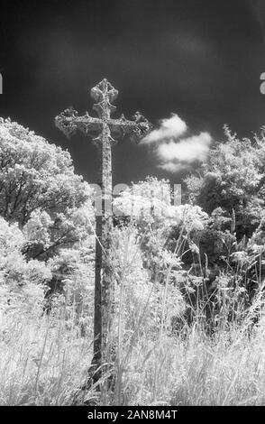 Stone cross marking a grave in the churchyard of Holy Trinity church, Merepond Lane, Privett, Alton, Hampshire, England, UK.  Black and white infra-red filmstock, with its characteristic prominent grain structure, high-contrast and glowing bright foliage: spooky, gothic atmosphere