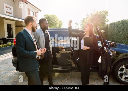 Side view photo of young female consultant with clipboard showing car keys to buyers, African and Caucasian men, showing a new car in auto show Stock Photo
