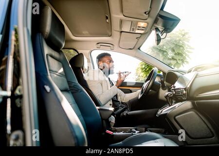 Young successful African American businessman talking on speakerphone through microphone with client, sitting in the expensive car. Negotiations and Stock Photo