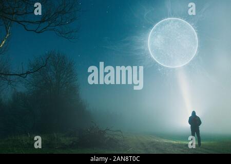 A man standing with a torch looking up at a glowing science fiction portal. On a foggy, winters night in the countryside. Stock Photo