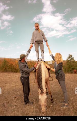 A teenage girl stands on a horse, supported on both sides by adults.