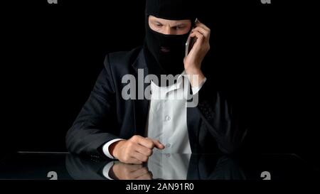 Murderer in mask talking on smartphone with crime partner, contract murder, gun Stock Photo