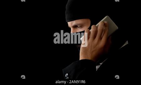 Killer in balaclava talking on phone, planning crime with partners, corruption Stock Photo
