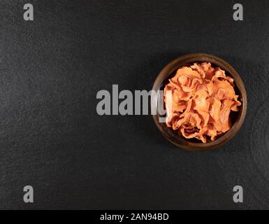 Dried carrot chips in a wooden bowl on a black stone background. Organic natural food. Top view, flat lay. Copy space. Stock Photo