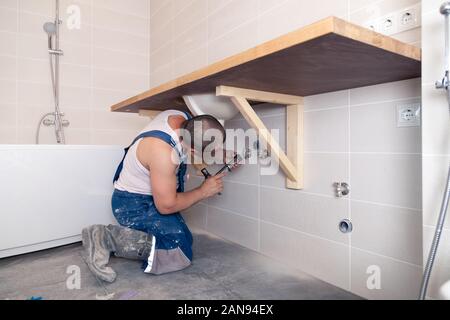 Closeup male plumber worker in blue denim uniform, overalls, fixing sink in bathroom with tile wall. Professional plumbing repair service, installatio Stock Photo
