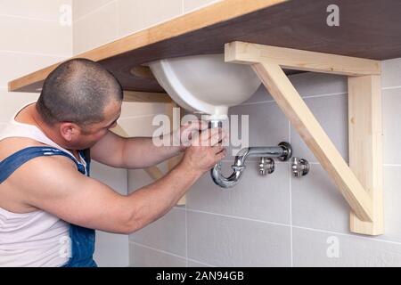 Closeup male plumber worker in blue denim uniform, overalls, fixing sink in bathroom with tile wall. Professional plumbing repair service, installatio Stock Photo