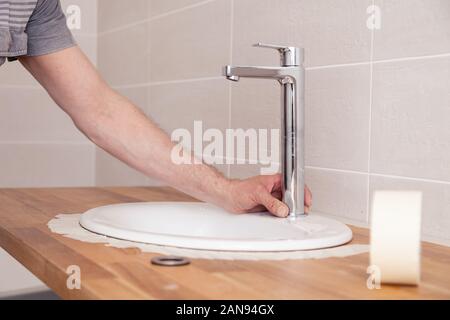 Closeup hands of a professional plumber worker installs a white oval ceramic sink on a wooden tabletop in the bathroom with beige tile, paste over the Stock Photo