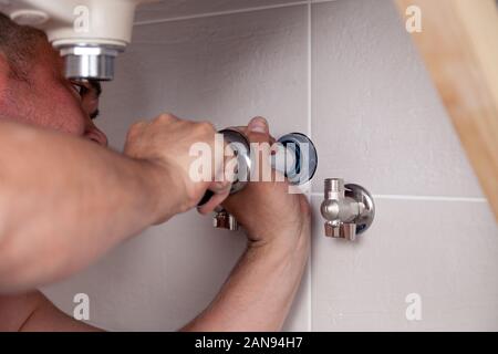 Closeup male plumber worker in blue denim uniform, overalls, fixing sink in bathroom with tile wall. Professional plumbing repair service, installatio Stock Photo