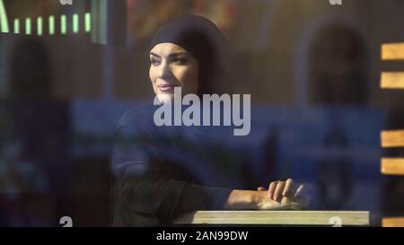 Smiling arab woman looking at city through window sitting in cafe, leisure Stock Photo