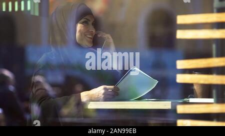 Young Muslim businesswoman in cafe holding documents, talking on phone, gadget Stock Photo