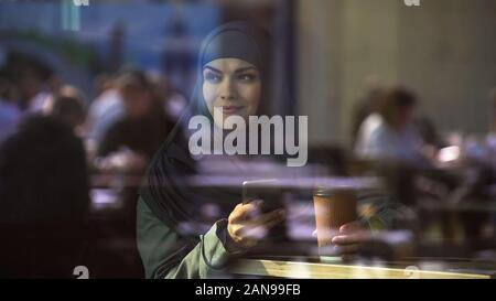 Smiling muslim woman with smartphone in hand looking through cafe window, city Stock Photo