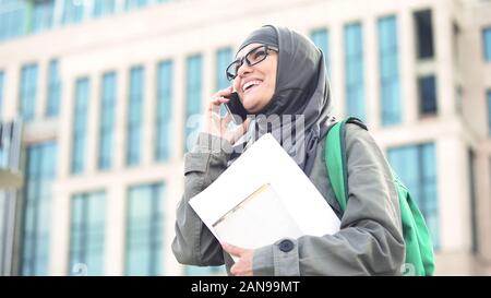 Cheerful Arabic female student talking on phone standing on university campus Stock Photo