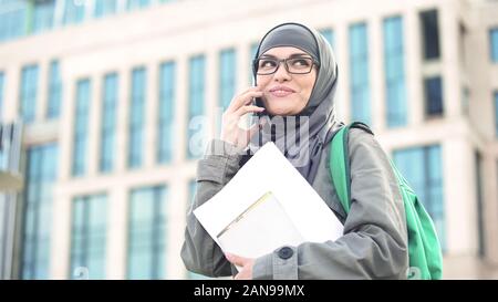 Happy lady in muslim hijab talking on phone, holding documents in hand, campus Stock Photo