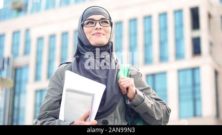 Inspired young female student wearing hijab smiling, standing outdoors on campus Stock Photo