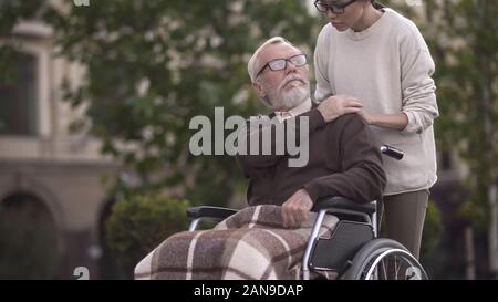 Aged sad disabled male in wheelchair covering young lady hand, family support Stock Photo