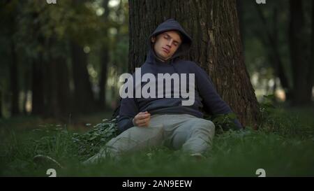 Vulnerable alcoholic sleeping under tree in park, careless and crazy youth Stock Photo