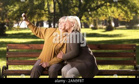Two old ladies taking selfie on mobile phone sitting on bench in park technology Stock Photo