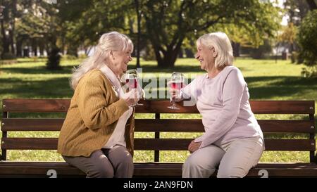 Two elderly women enjoying wine and chatting on bench in park happy golden years Stock Photo