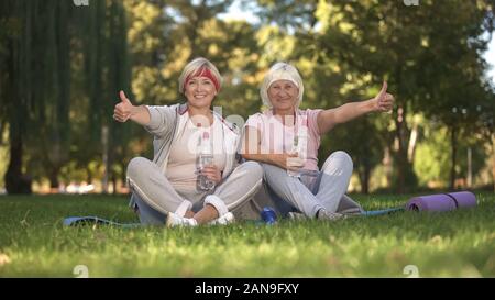 Two women showing thumbs up sitting on grass after doing exercises, positive Stock Photo