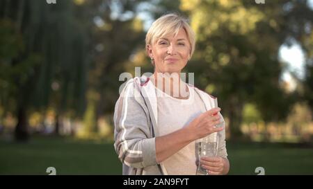 Pretty middle aged woman drinking water in park, keeping water balance, health Stock Photo