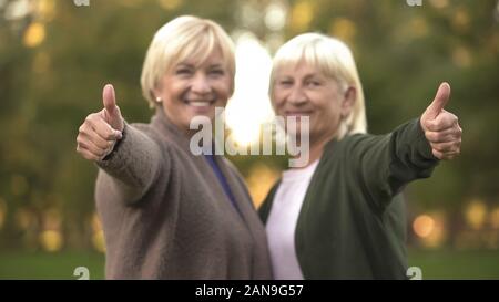 Smiling senior women showing thumbs up gesture, hugging and resting at park Stock Photo
