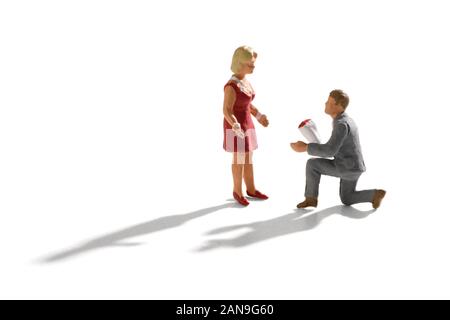 Miniature scene of engagement proposal, with a small man in grey suit, down on his knee with a bouquet of flowers, proposing to a woman in red dress. Stock Photo