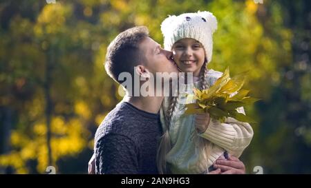 Dad holding daughter in funny hat and kissing, leisure together, autumn stroll Stock Photo