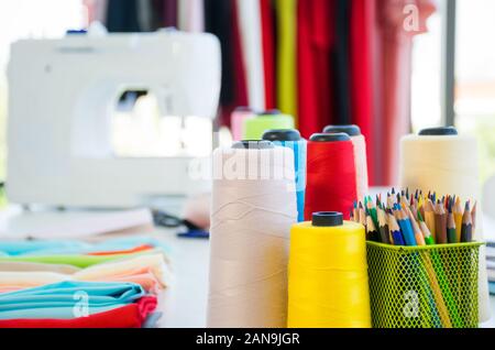 Front view of fashion designer working desk with colorful sewing thread, fabric and  colored pencils including a sewing machine and hanging clothes Stock Photo