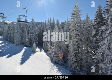 Empty chairs on cable going down, frozen fir forest and ski slope against sun with intended lens flare , view from ski lift Stock Photo