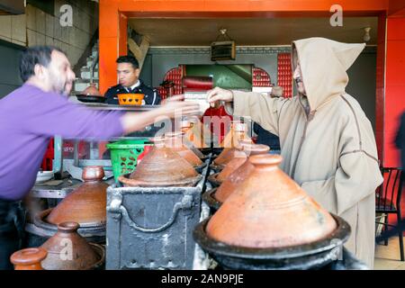 Marrakech, Morocco - January 7,2020: Berber man in traditional cloth with hood buying delicious tajine prepared and served in clay pots Stock Photo