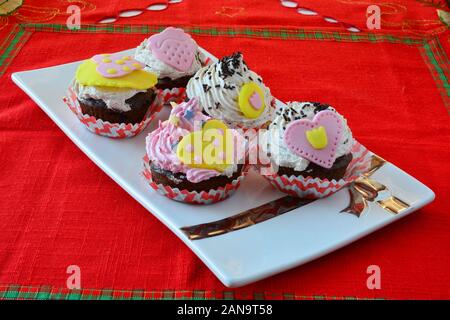 Love muffins, colorful velvet cupcakes decorated with marzipan hearts served in white ceramic saucer on table covered with red table cloth, side view Stock Photo