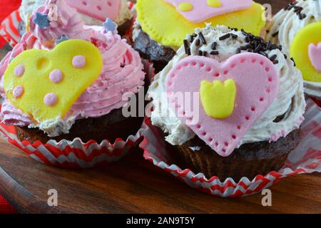 Valentine love muffins, colorful cupcakes decorated with marzipan hearts served on wooden round saucer over red table cloth, close up view Stock Photo