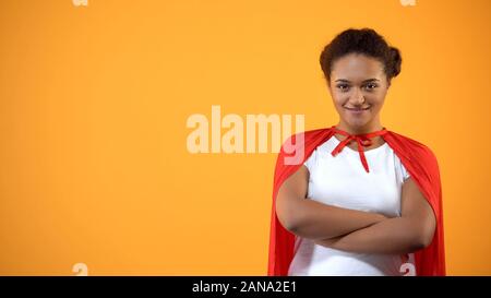 Confident young lady in red cape on bright background, powerful wonder woman Stock Photo