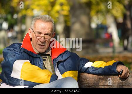 Good looking active senior holding cell phone and sitting on a bench in autumn park, conversation is over, enjoyment continues Stock Photo