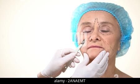 Surgeon placing scalpel to aging female patient face with marks, face lifting Stock Photo