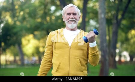 Handsome aged man doing arm exercises with dumbbells in park, leisure activity Stock Photo