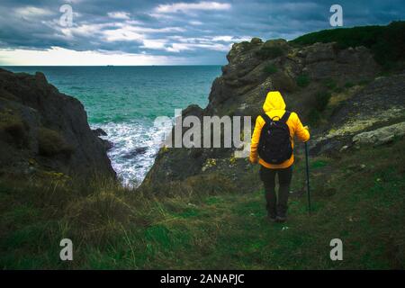 A Young man adventurer back shot with contrast orange jacket walking over path to sea among clifs with gloomy sky under dramatic clouds Stock Photo