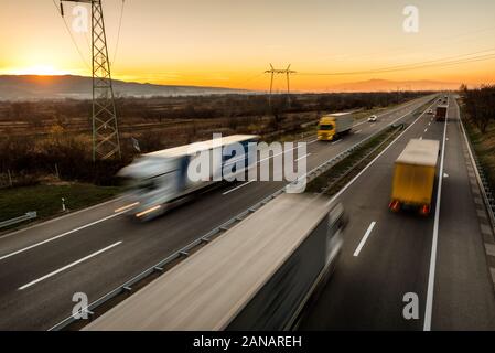 Delivery trucks and cars in high speed driving on a highway through rural landscape. Fast blurred motion drive on the freeway. Freight scene on the mo Stock Photo