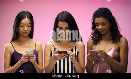 Female friends in pajamas chatting on smartphones, isolated on pink background Stock Photo