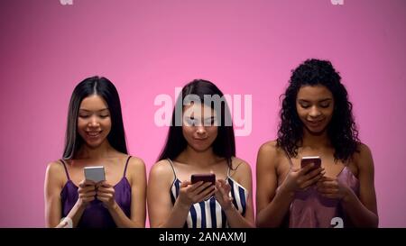 Three multiracial women scrolling smartphone on pink background, place for text Stock Photo