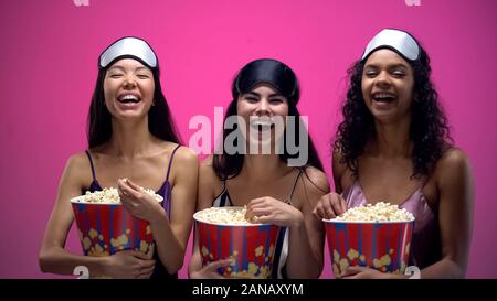 Laughing women in pajamas and eye masks watching funny comedy show with popcorn Stock Photo