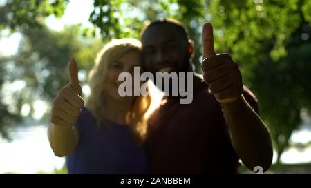 Smiling interracial couple showing thumbs up and looking at camera, relationship Stock Photo