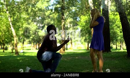 Surprise engagement in park, kneeling man makes proposal with ring, happy couple Stock Photo